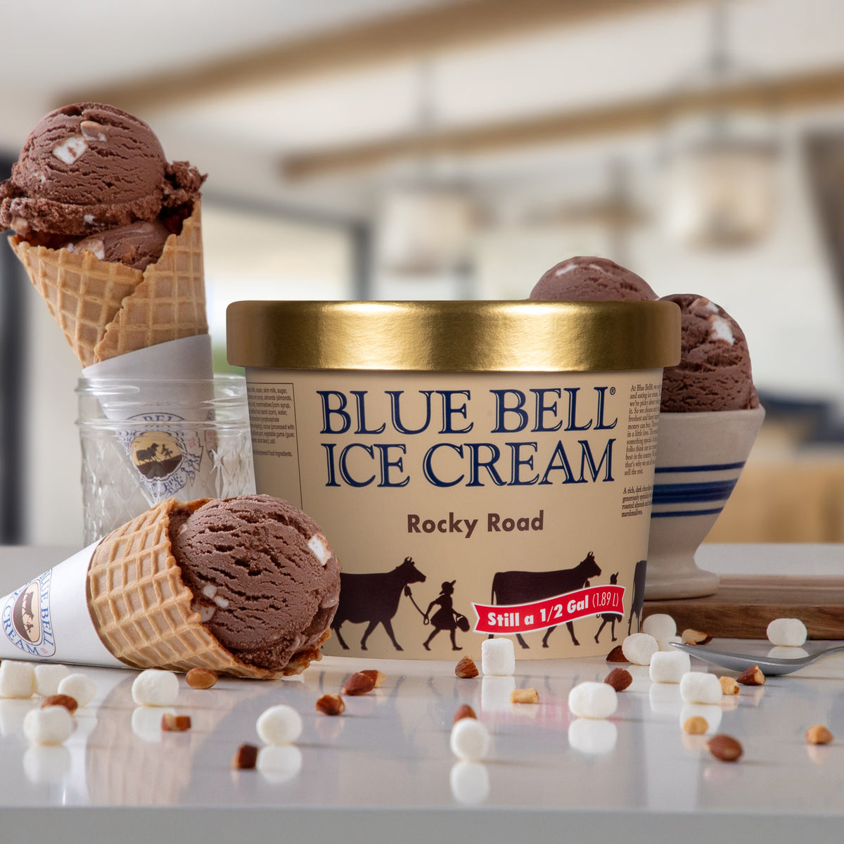 A tub of Blue Bell Rocky Road Ice Cream sits on a countertop with several chocolate ice cream scoops and cones arranged around it. The product label is beige with blue text, a gold lid, and black cow illustrations, while the surrounding ice cream cones and bowls add brown chocolate tones and small white marshmallow accents.