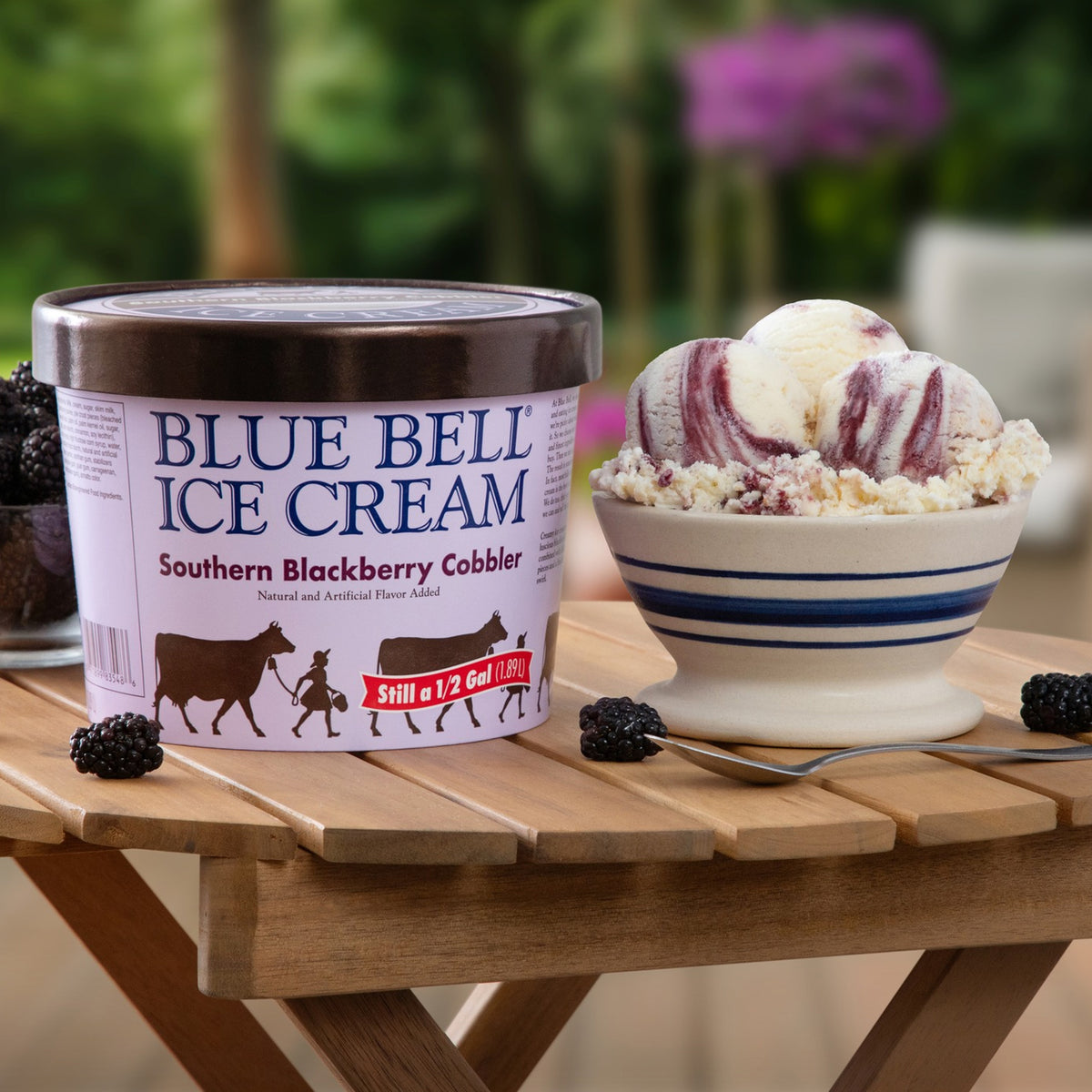 A tub of Blue Bell Ice Cream Southern Blackberry Cobbler flavor sits on a wooden table next to a bowl of the ice cream swirled with blackberry pie crust, with fresh blackberries and an outdoor garden blurred in the background.