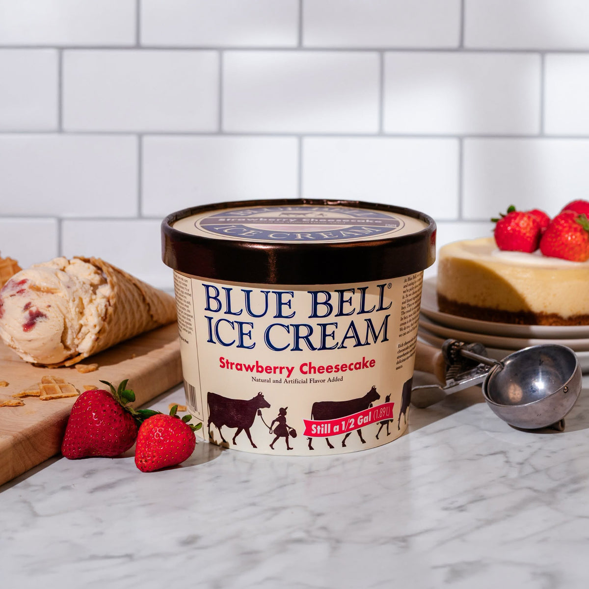 A tub of Blue Bell Strawberry Cheesecake Ice Cream with a waffle cone filled with strawberry cheesecake ice cream.  A strawberry cheesecake and dipper are in the background in a white kitchen setting. 