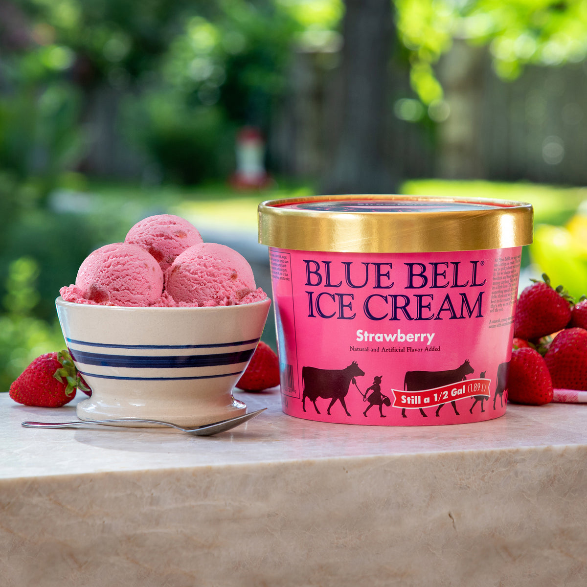 A tub of Blue Bell Ice Cream Strawberry flavor sits on a stone surface outdoors, with a bowl of pink ice cream scoops and fresh strawberries around it. The product tub is pink with a gold lid and blue branding text.
