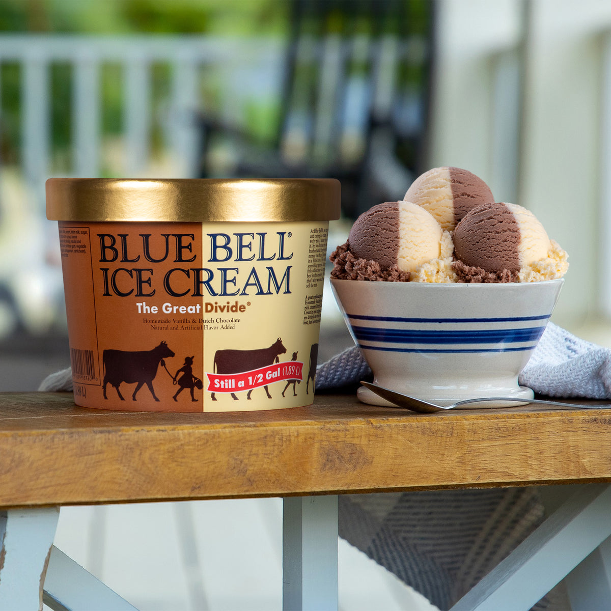 A tub of Blue Bell The Great Divide Ice Cream sits on a wooden table next to a bowl of scoops of ice cream in an outdoor porch setting.