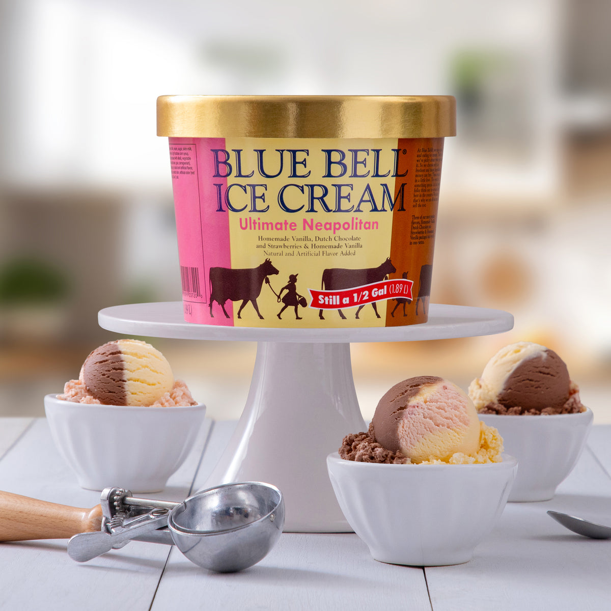 A tub of Blue Bell Ultimate Neapolitan Ice Cream on a white cake stand with white bowls and scoops of Neapolitan ice cream on a blurred background.