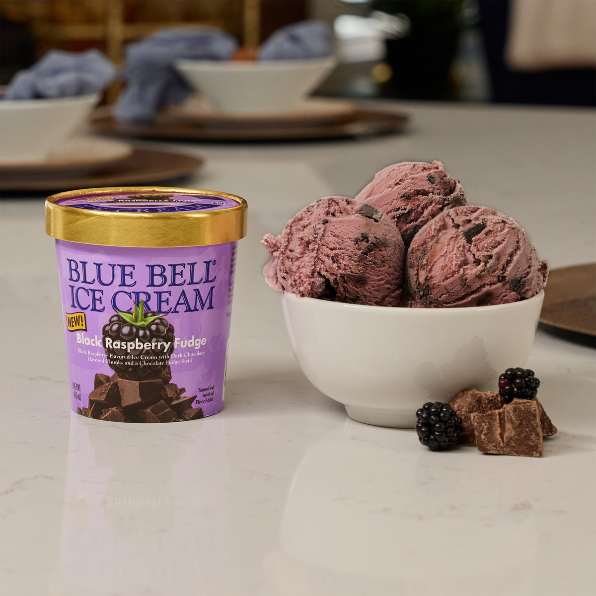 A pint of Blue Bell Ice Cream Raspberry Fudge flavor sits on a white marble countertop next to a white bowl filled with dark berry ice cream scoops, with a few black raspberries and chocolate chunks nearby.