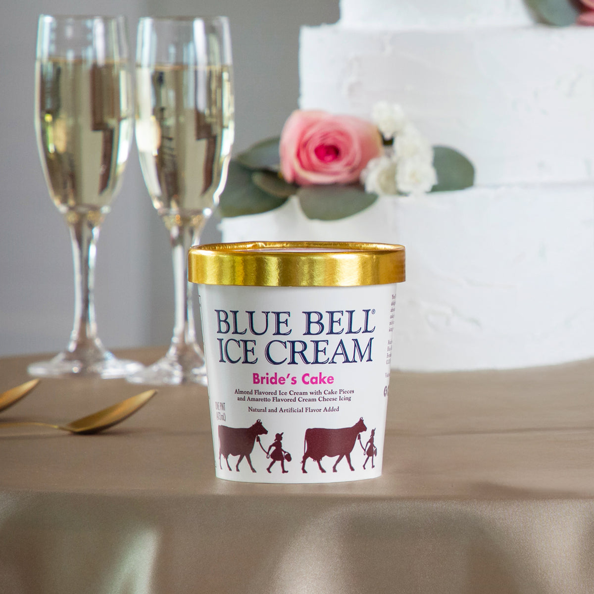 A pint of Blue Bell Ice Cream labeled 'Bride's Cake' sits on a table with champagne flutes and a white wedding cake blurred in the background.
