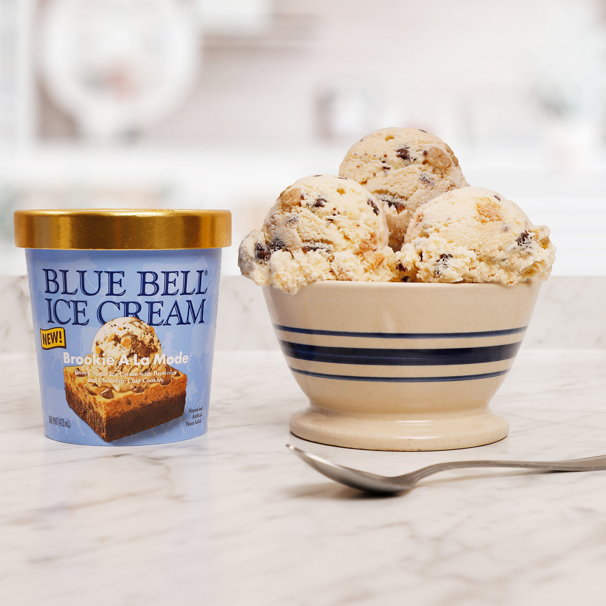 A tub of Blue Bell Brookie A La Mode Ice Cream next to a bowl filled with scoops of  ice cream on a light marble surface.