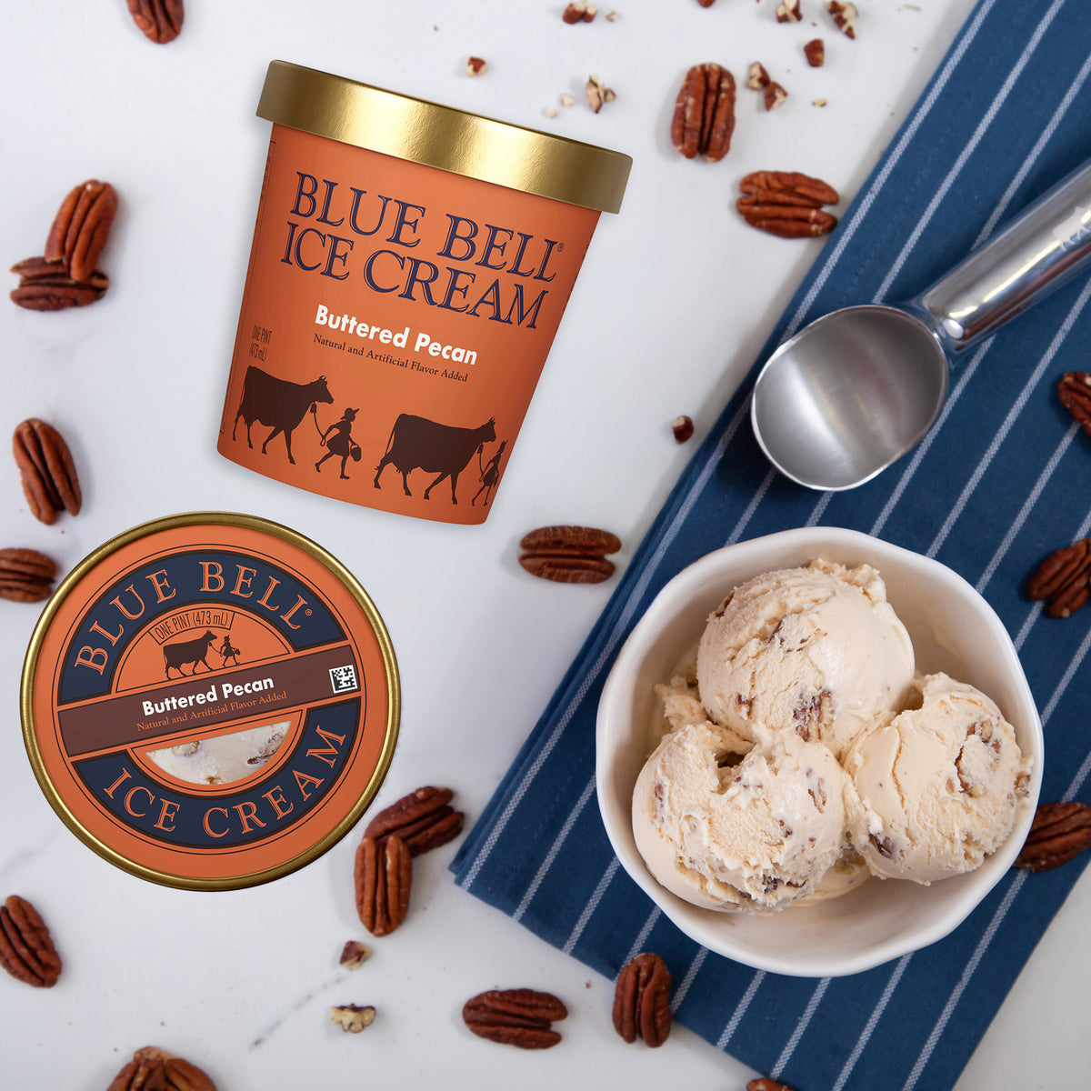 Blue Bell Ice Cream Buttered Pecan flavor shown as an orange-labeled tub, a matching lid, and scoops of the cream in a bowl on a blue striped napkin, with scattered pecans on a white surface.