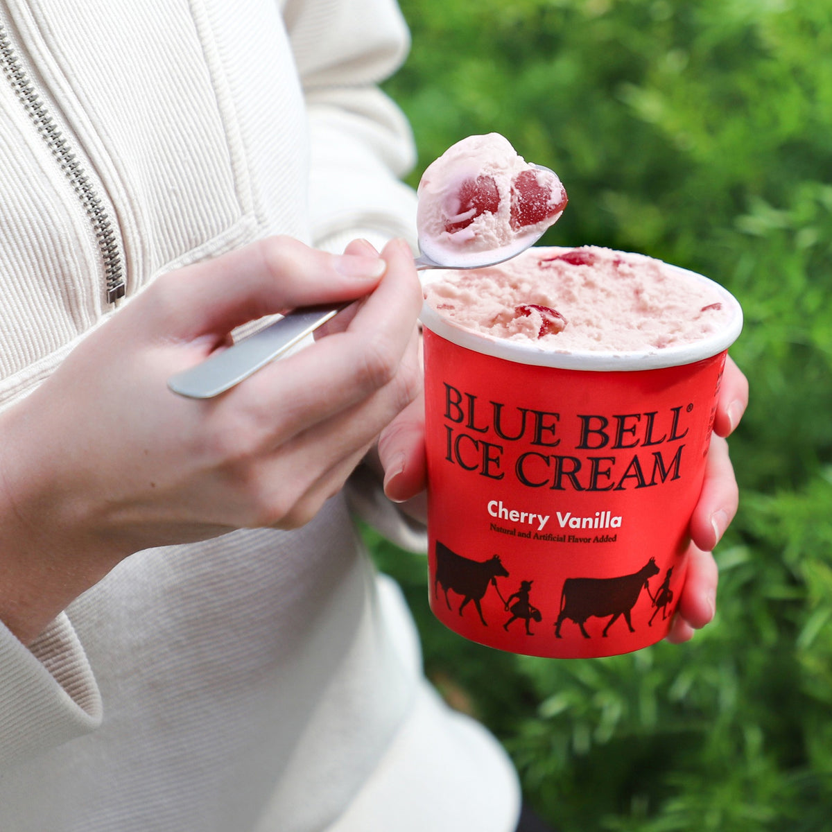 A person in a cream zip-up jacket holds a red cup of Blue Bell Cherry Vanilla Ice Cream in their other hand, with a spoonful of the pinkish vanilla-cherry ice cream resting above the cup. The background is a soft-focus green outdoor setting.