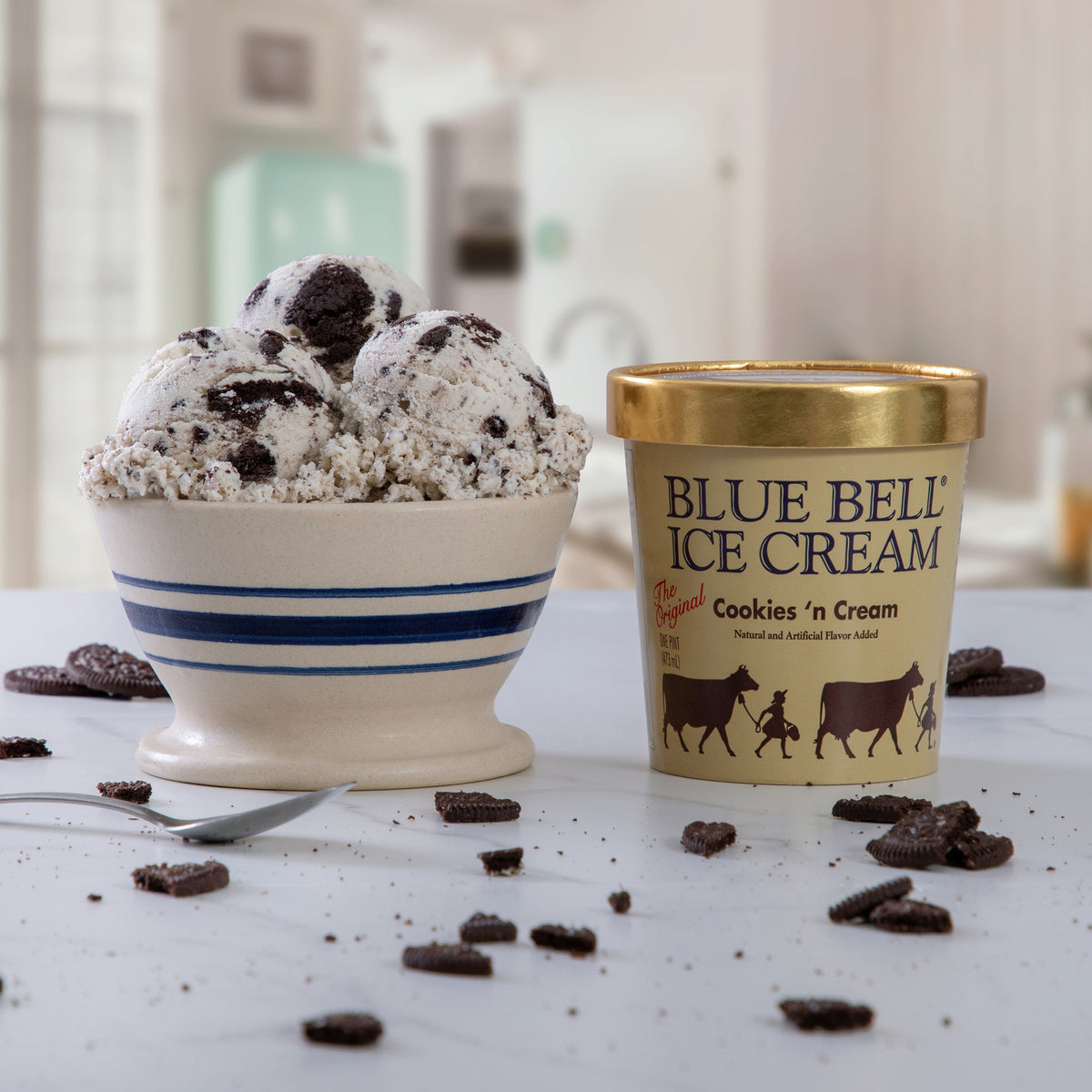 A bowl of Blue Bell Ice Cream Cookies 'n Cream flavor with cookie crumb toppings sits next to its branded pint container on a light countertop, with scattered cookie pieces and a spoon around it.