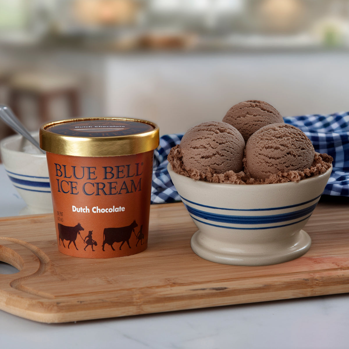 A pint of Blue Bell Ice Cream Dutch Chocolate ice cream sits on a wooden cutting board next to a bowl filled with chocolate ice cream scoops, with a blurred kitchen background.