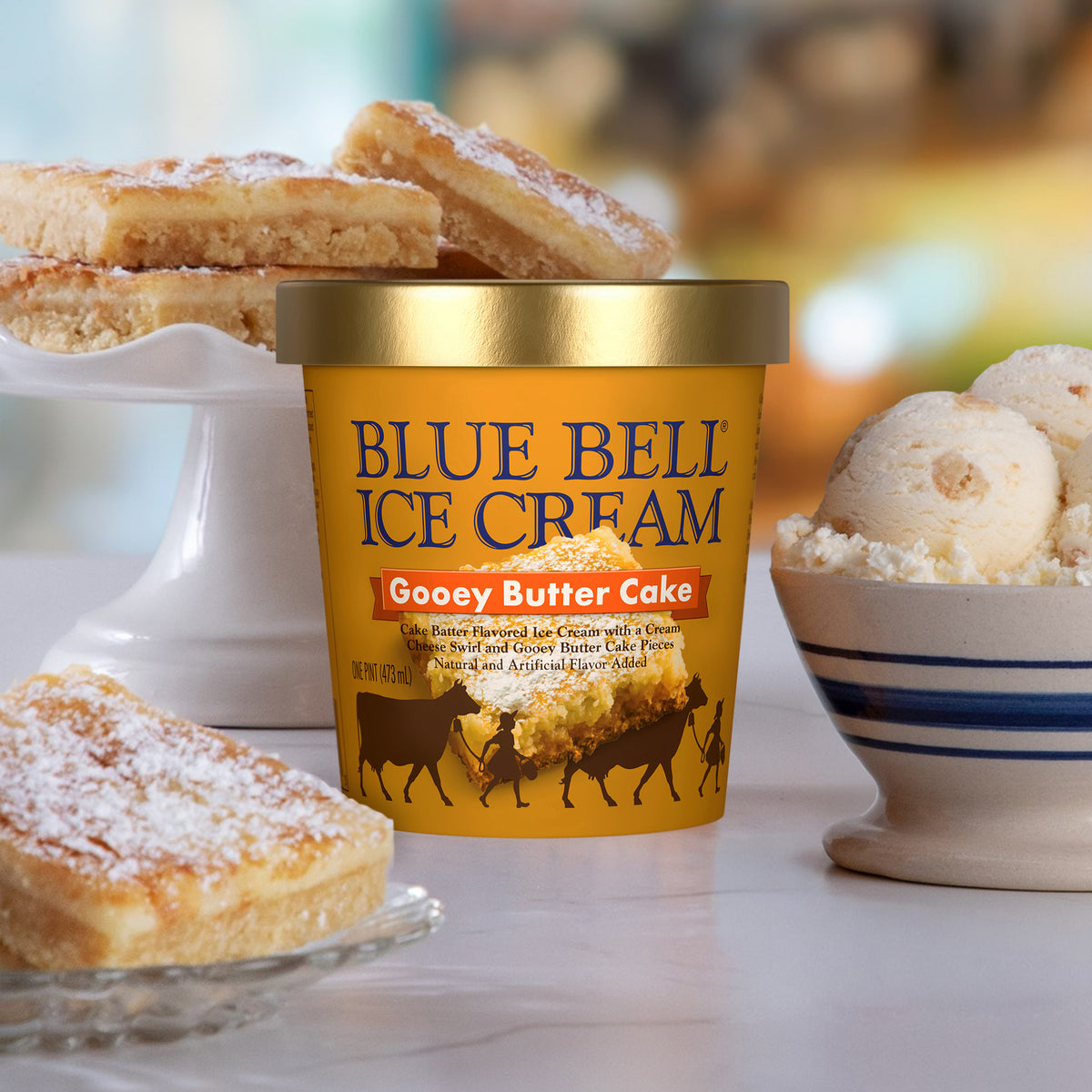 A tub of Blue Bell Ice Cream in Gooey Butter Cake flavor stands on a countertop, with butter cake slices on a pedestal and a bowl of scoops of ice cream in a softly blurred kitchen setting.