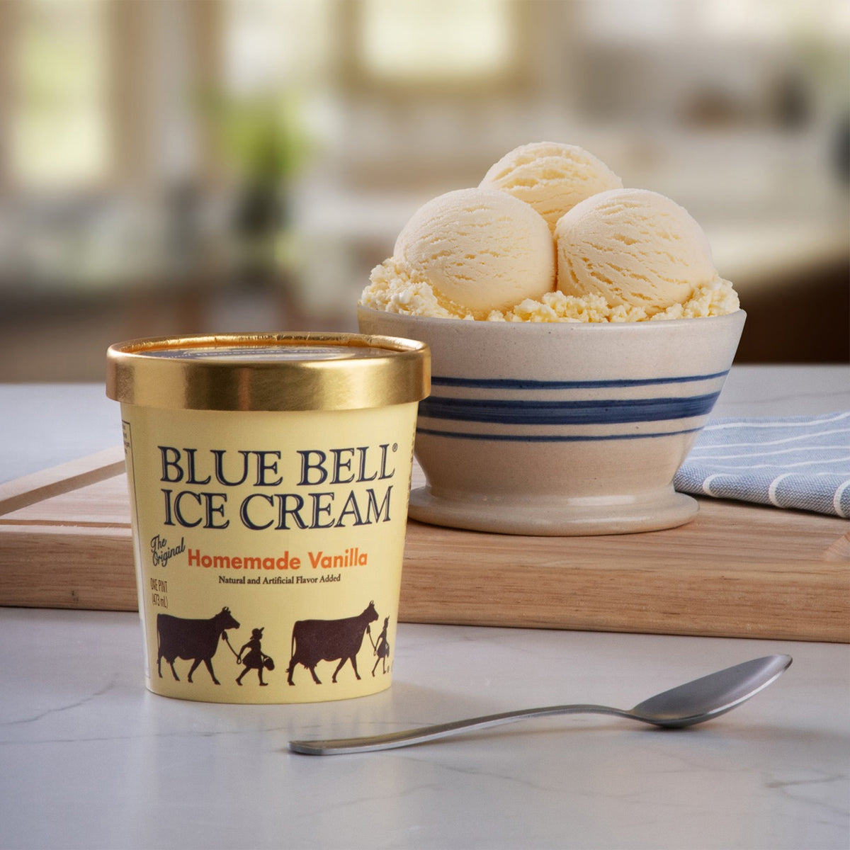 A pint of Blue Bell Ice Cream Homemade Vanilla sits on a wooden board on a marble countertop, with a bowl of vanilla ice cream scoops and a spoon in a softly blurred kitchen setting.