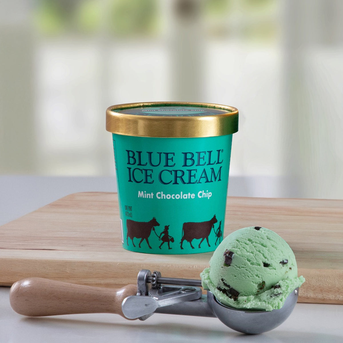 A pint of Blue Bell Ice Cream in Mint Chocolate Chip flavor sits on a wooden board with a metal ice cream scoop holding a scoop of mint green ice cream in the foreground.