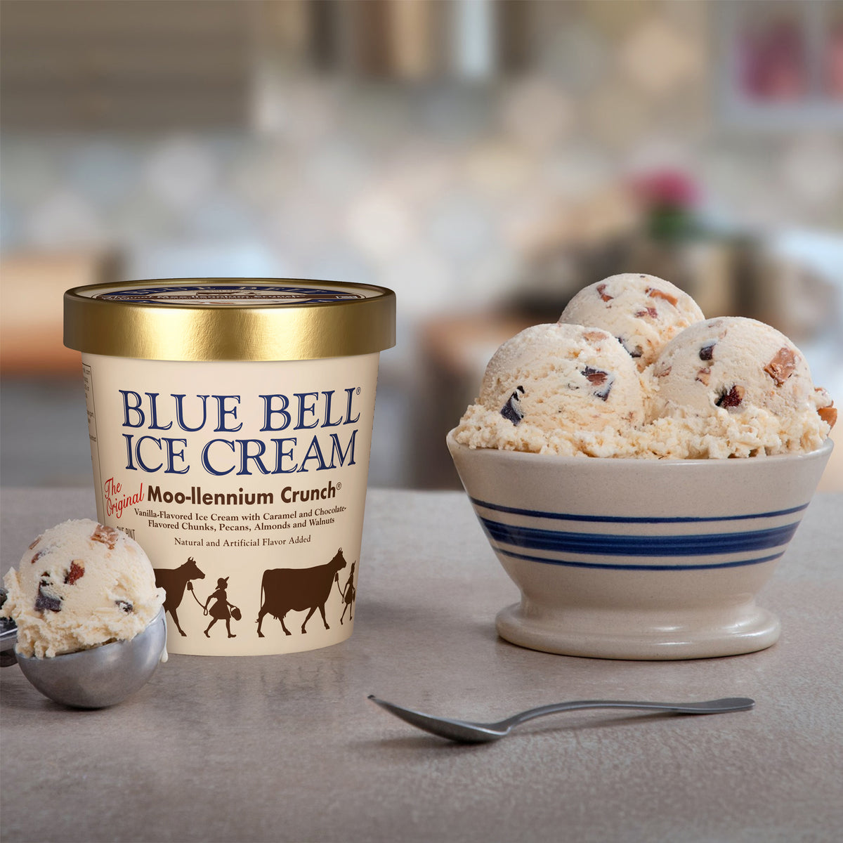 A pint of Blue Bell Ice Cream next to a bowl filled with scoops of Moo-llennium Crunch ice cream, with a spoon and another scoop in a softly blurred kitchen setting.