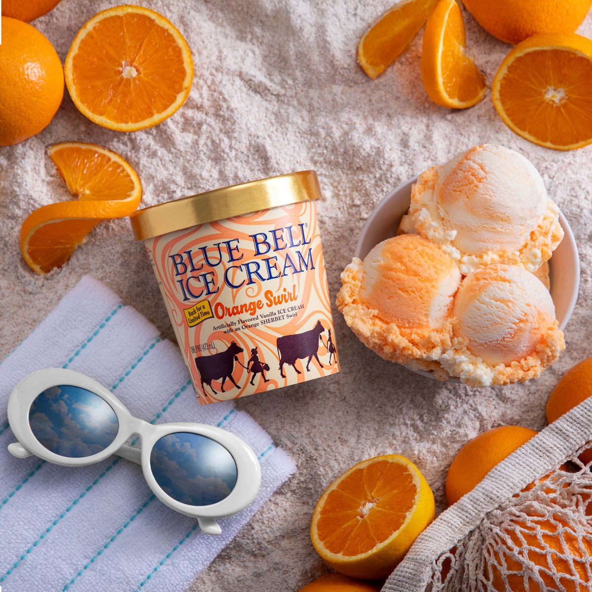 A tub of Blue Bell Ice Cream Orange Swril ice cream is styled in a beach-themed flat lay with fresh orange slices, white sunglasses on a striped towel, and scoops of the orange swirl ice cream in a bowl.