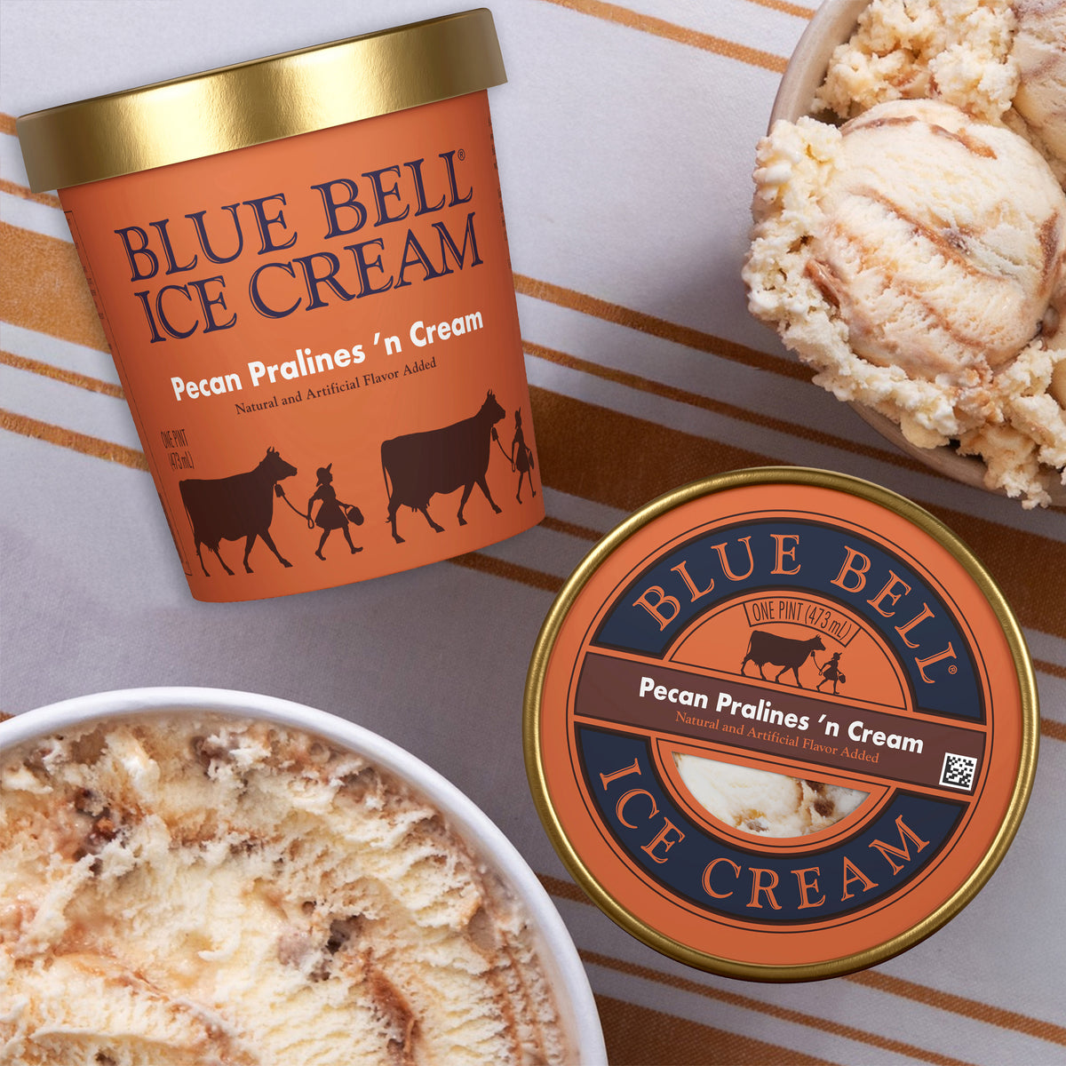 Two Blue Bell Ice Cream pint containers and bowls of Pecan Pralines 'n Cream ice cream arranged on a striped surface. The containers are orange with gold lids and dark blue branding, and the ice cream is light tan with pecan pieces and praline sauce swirl.