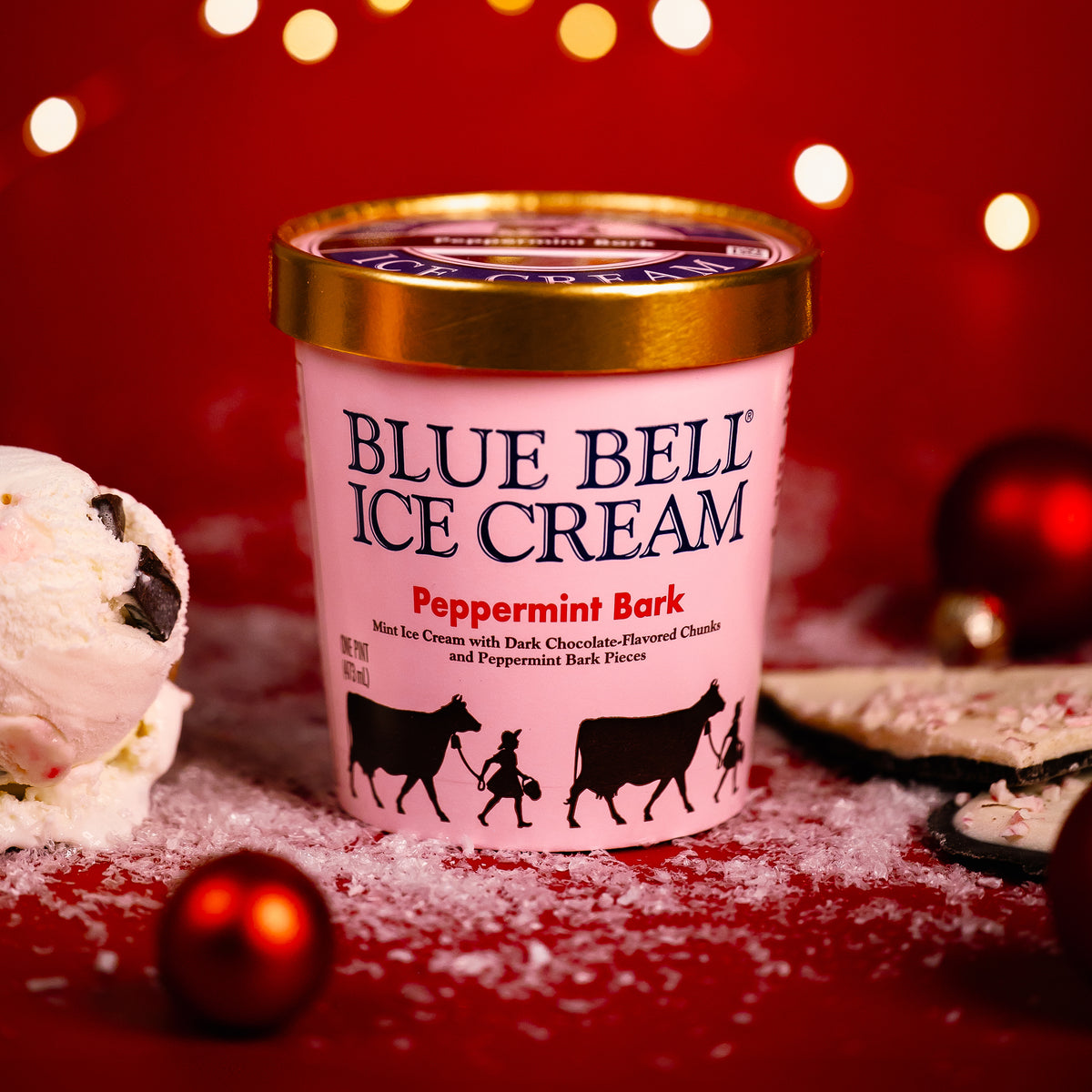 A pint of Blue Bell Ice Cream Peppermint Bark mint ice cream is centered on a red holiday-themed background with festive lights, candy canes, and cookie props. The container is pink with a gold lid and black cow illustrations on the front.