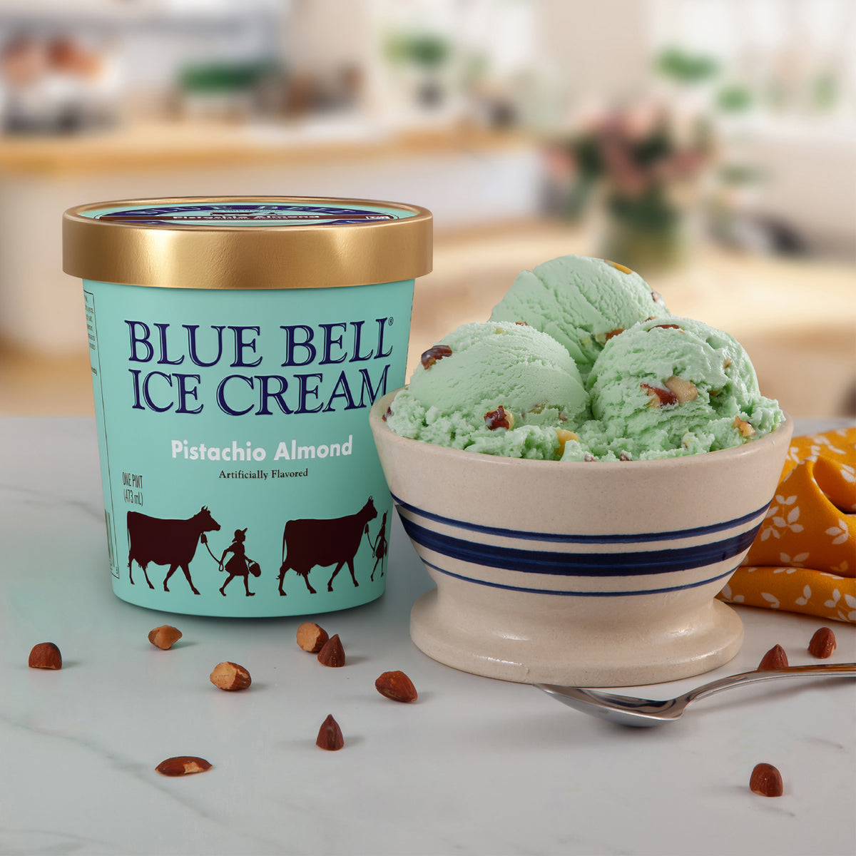 A pint of Blue Bell Ice Cream Pistachio Almond flavor stands next to a bowl of mint green pistachio ice cream with nuts, on a light marble surface with scattered almond pieces.