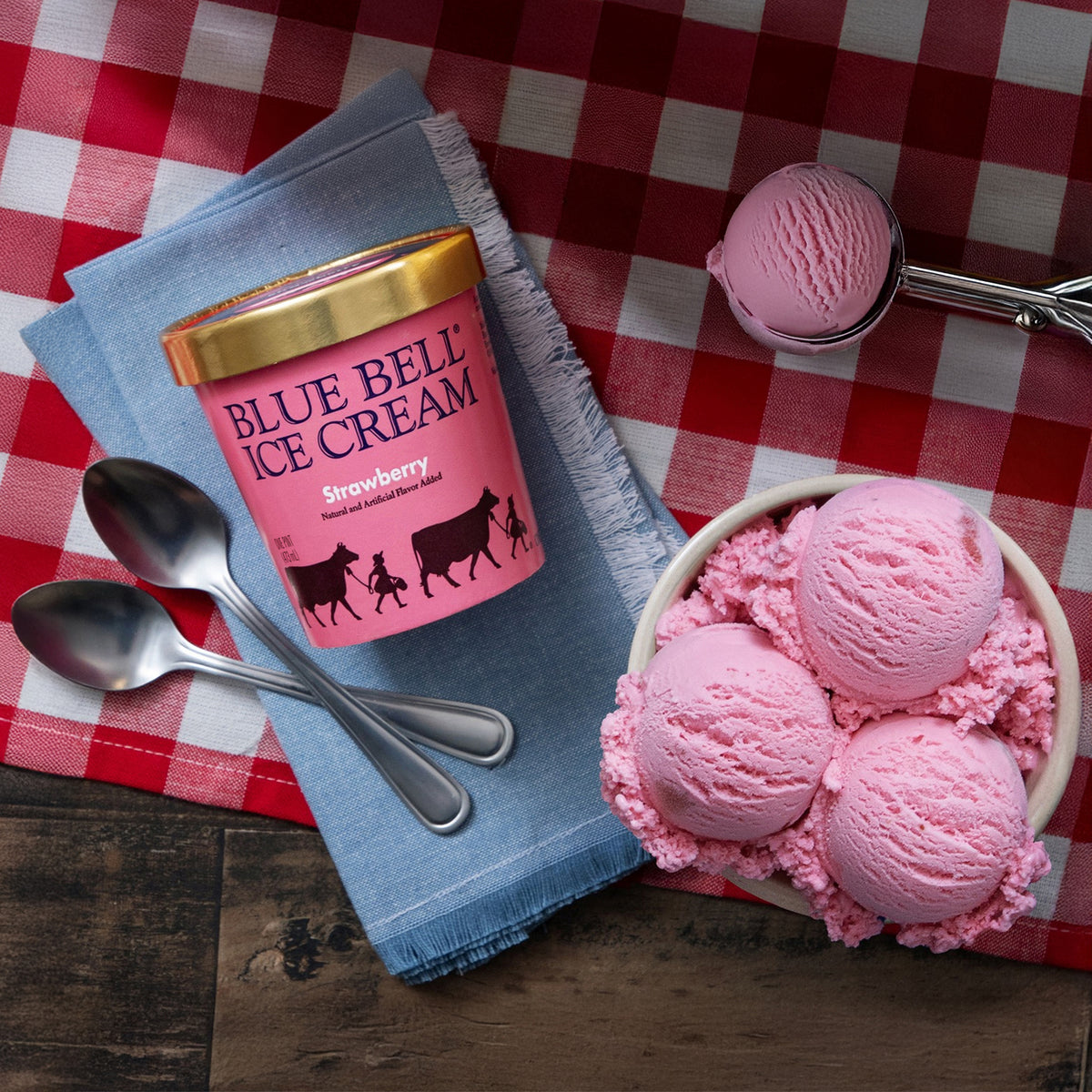 A pint of Blue Bell Ice Cream strawberry ice cream sits on a blue napkin beside two silver spoons, with a bowl of pink scoops of ice cream and a single scoop resting on a red-and-white checkered tablecloth over a wooden surface.