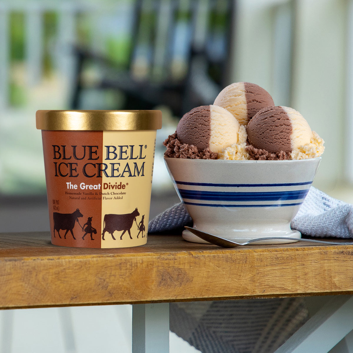 A pint of Blue Bell Ice Cream labeled 'The Great Divide' sits on a wooden table beside a bowl filled with scoops of the ice cream, with a blurred outdoor patio background.