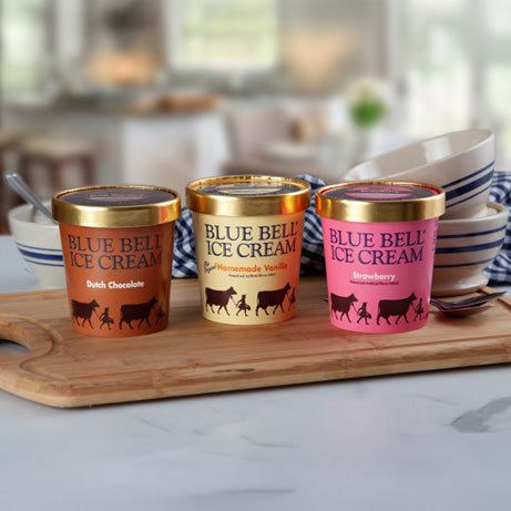 Three pints of Blue Bell Ice Cream on a wooden board with a kitchen background.